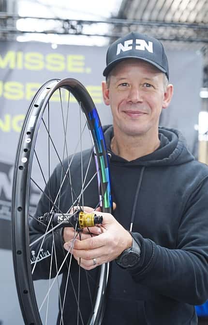 Eine Person hält ein Fahrrad-Laufrad mit Nabenaufdruck und bunter Felge, im Hintergrund Messestand.