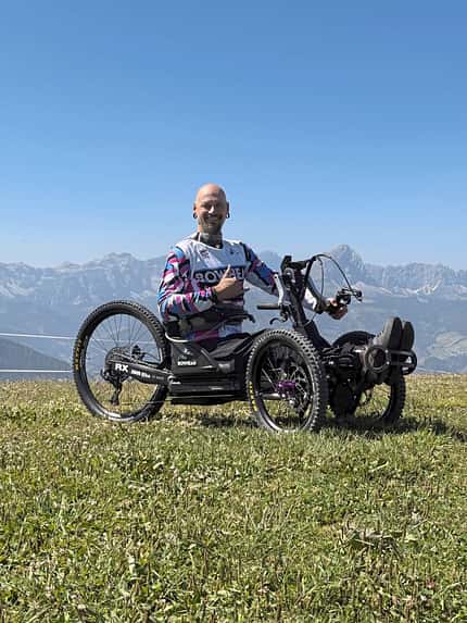 Eine Person sitzt in einem geländegängigen Handbike (Bowhead RX) auf einer Wiese vor Bergpanorama.